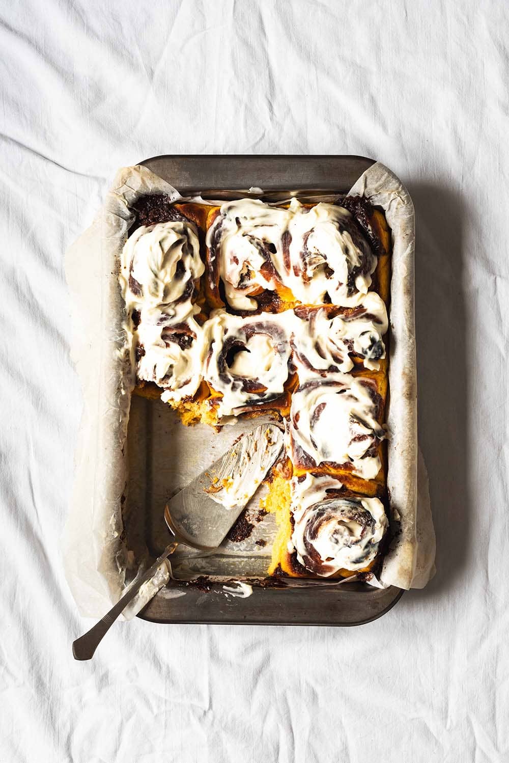 En una mesa, cubierta con mantel blanco, hay un molde rectangular. Los Cinnamon Rolls de calabaza están cubiertos con queso crema. En el molde está una pala para tartas. Ya faltan un par de trozos de Cinnamon Rolls del molde.