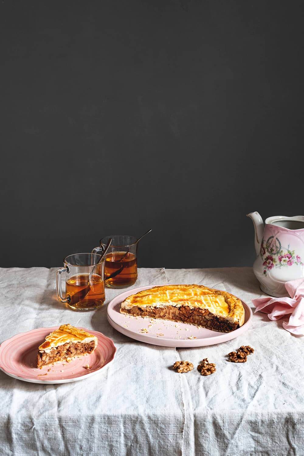 En una mesa, cubierta con mantel blanco, en el centro está la tarta de nueces Engadin. Alrededor hay dos tazas de té, una servilleta rosa y una tetera vintage. También al lado de la tarta Engadin hay un trozo de tarta ya cortada. Se ven bien jugoso. 