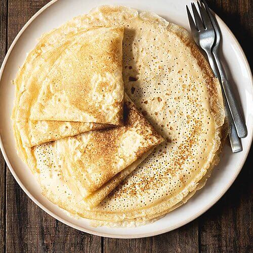 En una mesa de madera hay un plato blanco. En el plato hay muchas crepes caseras sin mantequilla. Al lado de las crepes hay dos tenedores. 