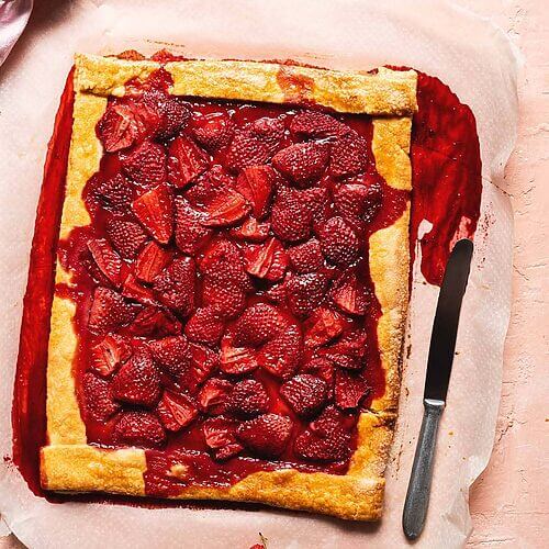 En una mesa de color rosa está un papel de horno. En el papel está el galette de fresas entero. Al lado, hay un cuchillo y una servilleta de color rosa y varias fresas silvestres.