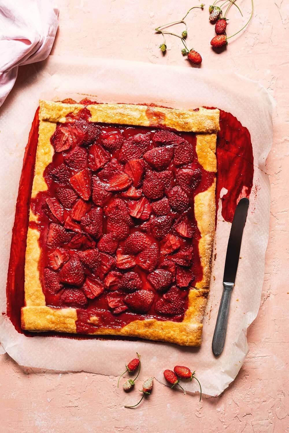 En una mesa de color rosa está un papel de horno. En el papel está el galette de fresas entero. Al lado, hay un cuchillo y una servilleta de color rosa y varias fresas silvestres.