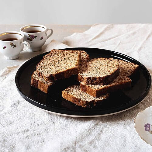 En una mesa cubierta con un mantel blanco hay un plato negro. En el plato hay varias rodajas de pan de especias. Al lado del plato hay dos tazas de té.