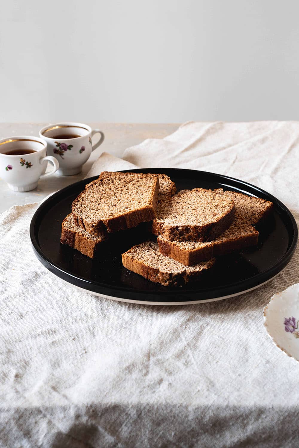 En una mesa cubierta con un mantel blanco hay un plato negro. En el plato hay varias rodajas de pan de especias. Al lado del plato hay dos tazas de té.