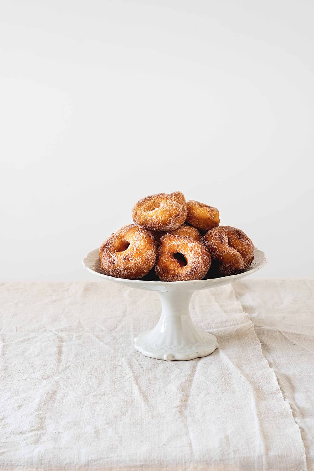 En una mesa hay un mantel blanco. En la mesa hay una tartera cerámica vintage. En esta tartera hay varias rosquillas de limón caseras y esponjosas.