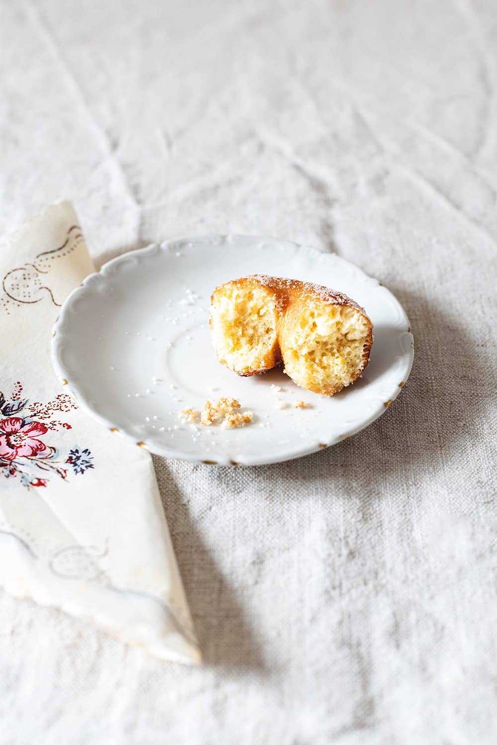 En una mesa hay un mantel blanco. En la mesa hay un plato blanco y en el plato hay una rosquilla de limón casera y está mordida. Se ve muy esponjoso. Al lado del plato hay una servilleta vintage.