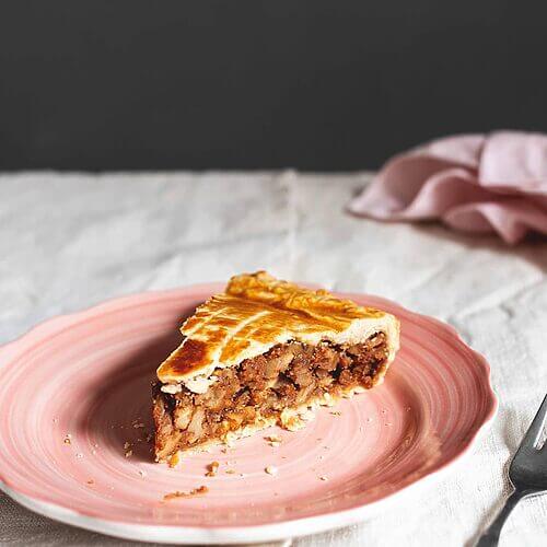 En una mesa, cubierta con mantel blanco está un plato rosa. En el plato hay un trozo de tarta de nueces Engadin. Se ve muy jugoso. Al lado del plato hay una servilleta rosa y un tenedor.