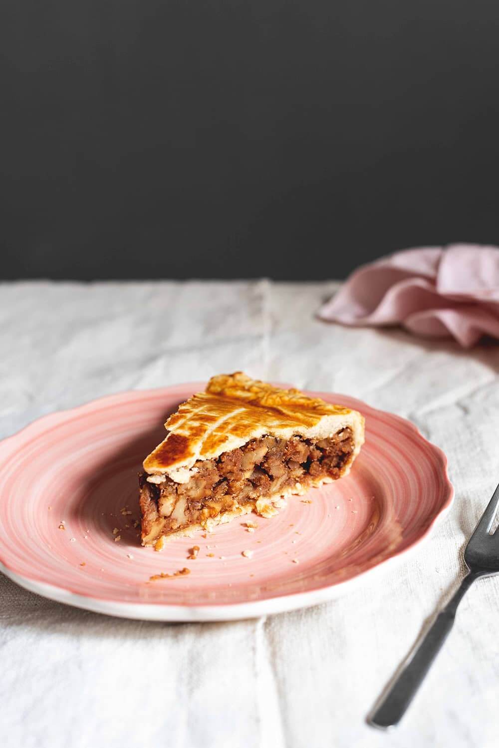 En una mesa, cubierta con mantel blanco está un plato rosa. En el plato hay un trozo de tarta de nueces Engadin. Se ve muy jugoso. Al lado del plato hay una servilleta rosa y un tenedor.