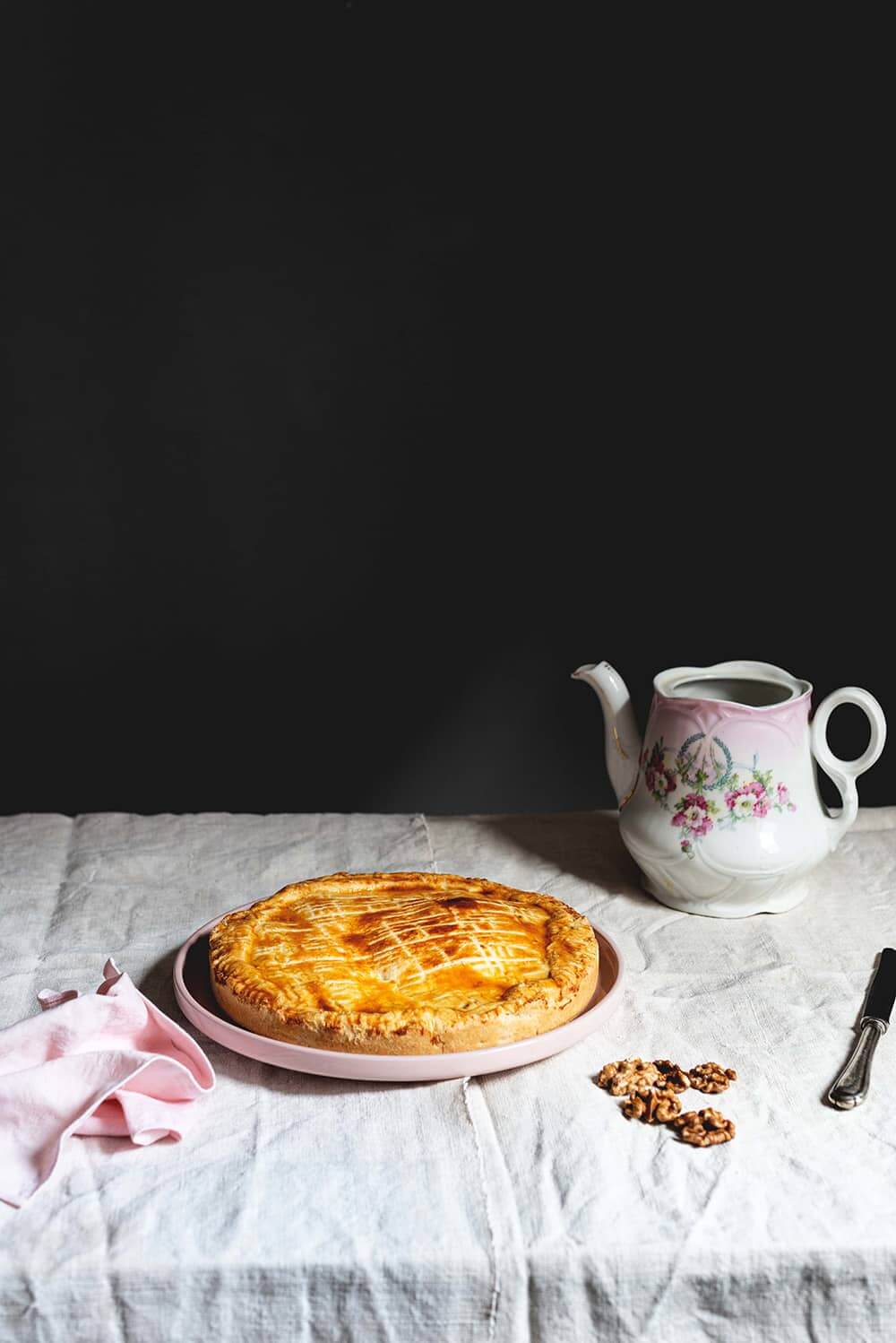 En una mesa, cubierta con mantel blanco, en el centro está la tarta de nueces Engadin. Alrededor hay dos tazas de té, una servilleta rosa, un cuchillo y una tetera vintage. La tarta no está cortada.