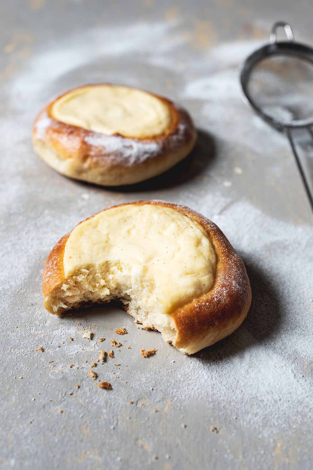 En una superficie gris hay dos Vatrushkas. La primera Vatrushka está mordida. Se ve muy esponjosa. Al lado de los bollos rellenos con queso crema hay una colador para espolvorearlas con azúcar glas. 