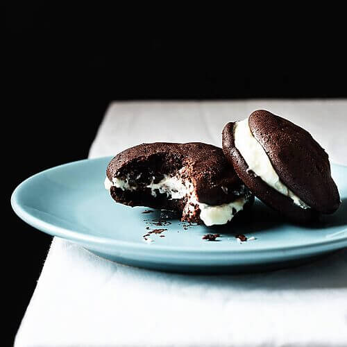 En una mesa hay un mantel blanco. En la mesa en un plato azul hay dos Whoopie pies de chocolate. Uno de ellos está mordido.