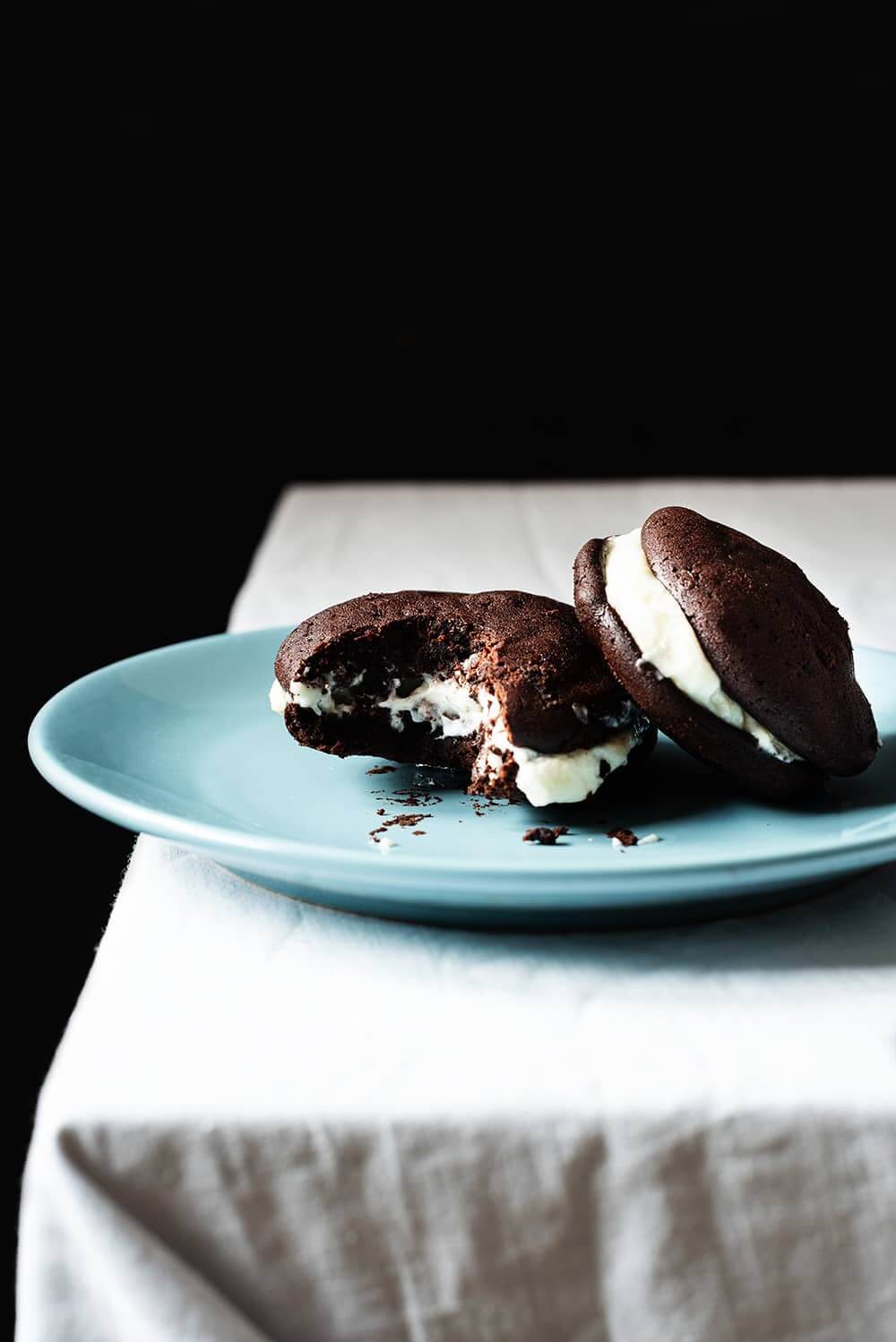 En una mesa hay un mantel blanco. En la mesa en un plato azul hay dos Whoopie pie de chocolate. Uno de ellos está mordido.