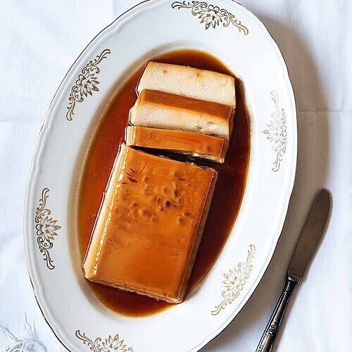 En una mesa está puesto una mantel blanca. En la mesa hay un cuenco blanco dónde está el flan de turrón sin horno con cuajada. El flan está cortado. Se ve cremoso. Al lado del cuenco hay un cuchillo.