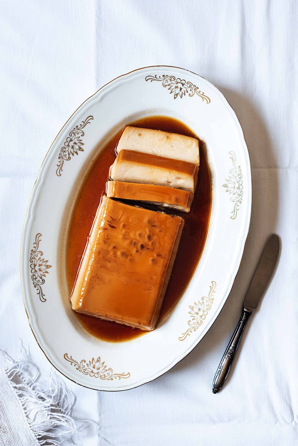 En una mesa está puesto una mantel blanca. En la mesa hay un cuenco blanco dónde está el flan de turrón sin horno con cuajada. El flan está cortado. Se ve cremoso. Al lado del cuenco hay un cuchillo.