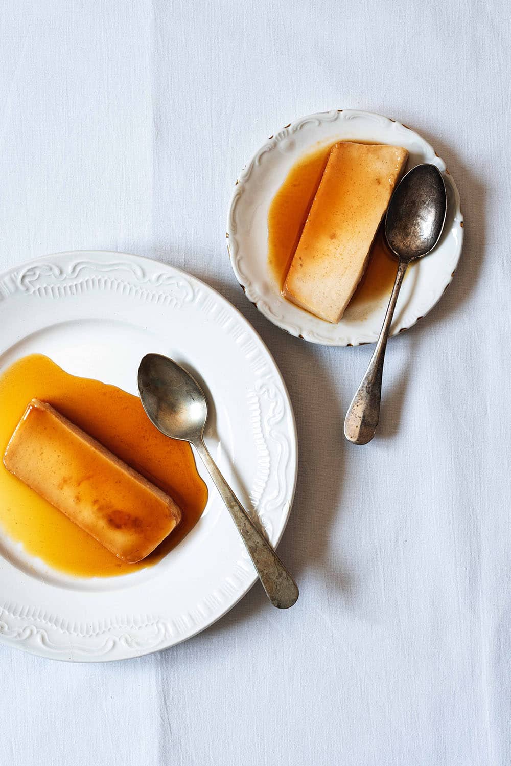 En una mesa está puesto una mantel blanca. En la mesa hay dos platos blancos. En cada plato hay un trozo de flan de turrón sin horno con cuajada y una cuchara.