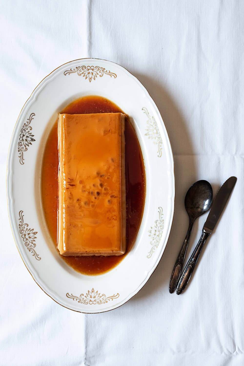 En una mesa está puesto una mantel blanca. En la mesa hay un cuenco blanco dónde está el flan de turrón sin horno con cuajada. Al lado del cuenco hay un cuchillo y una cuchara.