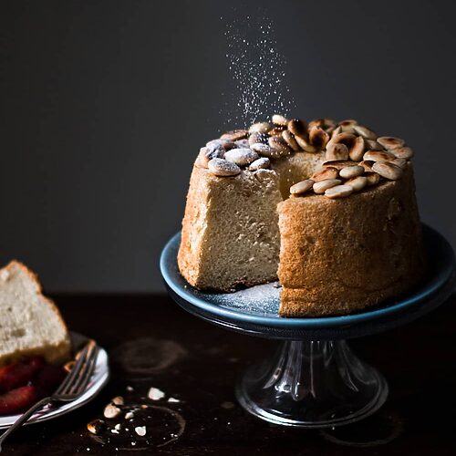 En una mesa de madera hay un porta tartas de cristal. En el porta tartas está el Angel Food Cake con almendra esponjoso. Una mano está espolvoreando con azúcar glas. Al lado del bizcocho hay una ración. 