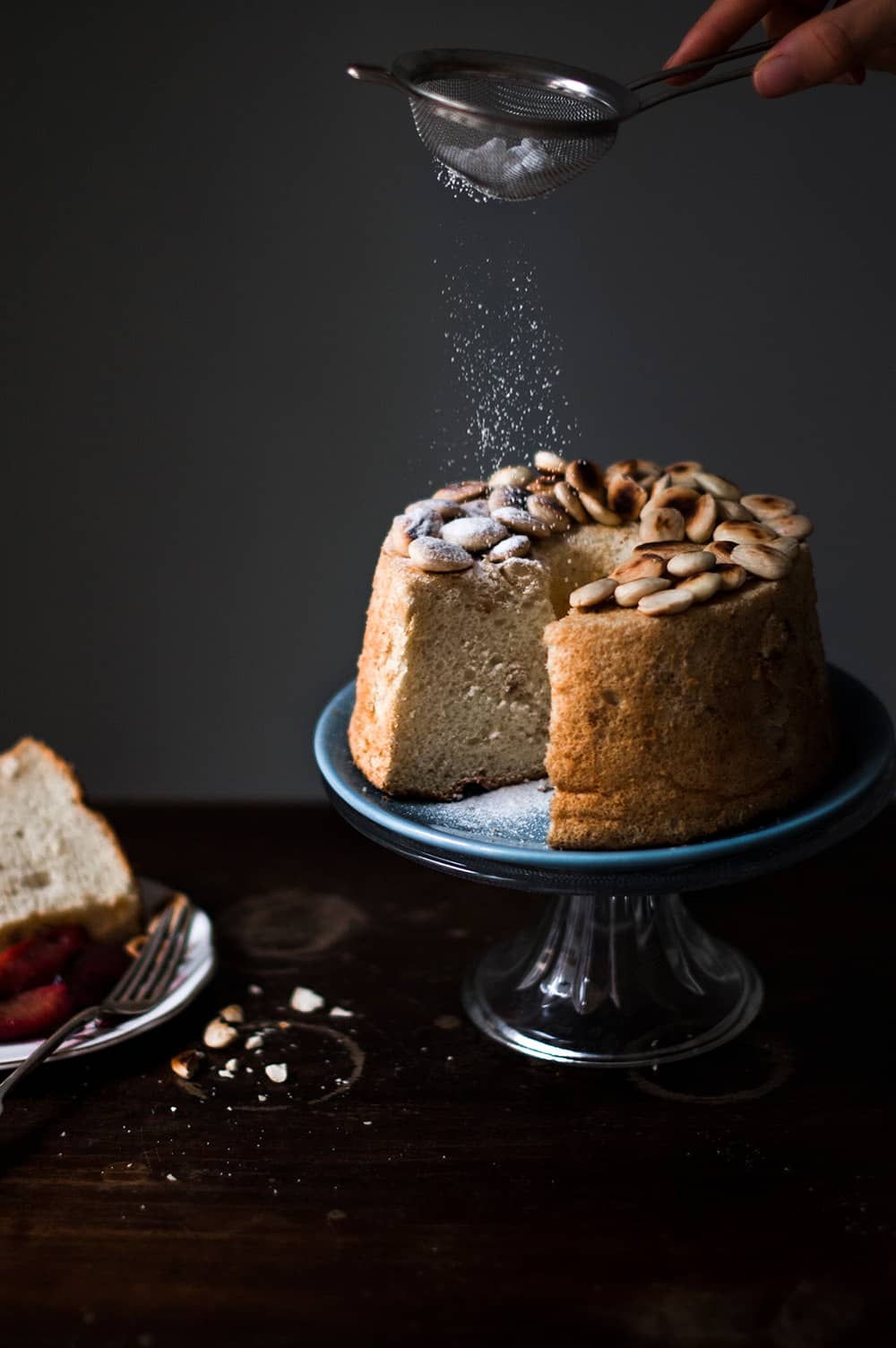 En una mesa de madera hay un porta tartas de cristal. En el porta tartas está el Angel Food Cake con almendra esponjoso. Una mano está espolvoreando con azúcar glas. Al lado del bizcocho hay una ración. 