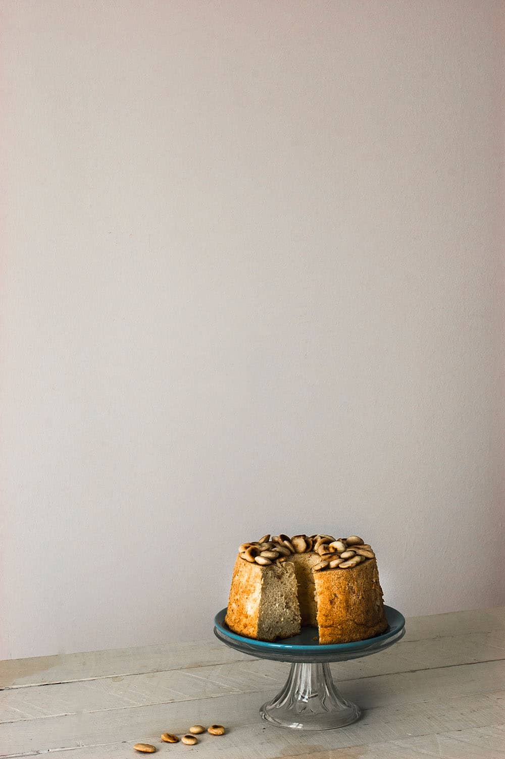 En una mesa de madera blanca hay un porta tartas de cristal. En el porta tartas está el Angel Food Cake con almendra esponjoso. Al lado del bizcocho hay almendras tostadas