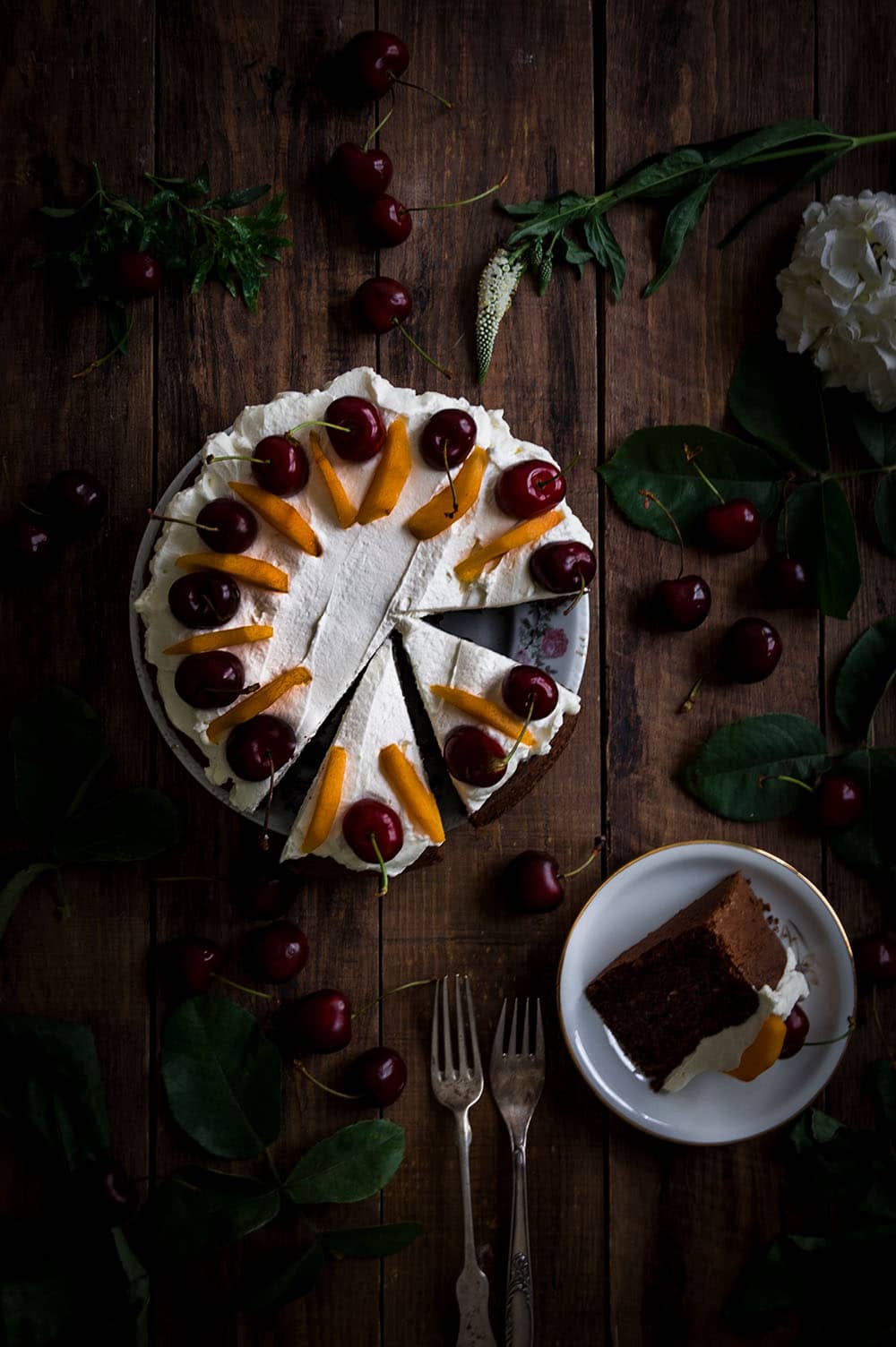 En una mesa de madera hay un plato. Encima está el bizcocho de chocolate y nueces sin gluten. Está cubierto con nata montada y decorado con frutas frescas. Al lado del bizcocho hay una ración.