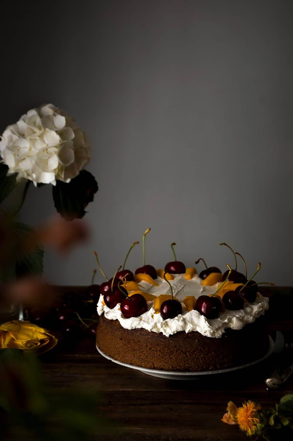 En una mesa de madera hay un plato. Encima está el bizcocho de chocolate y nueces sin harina. Alrededor hay flores frescas.