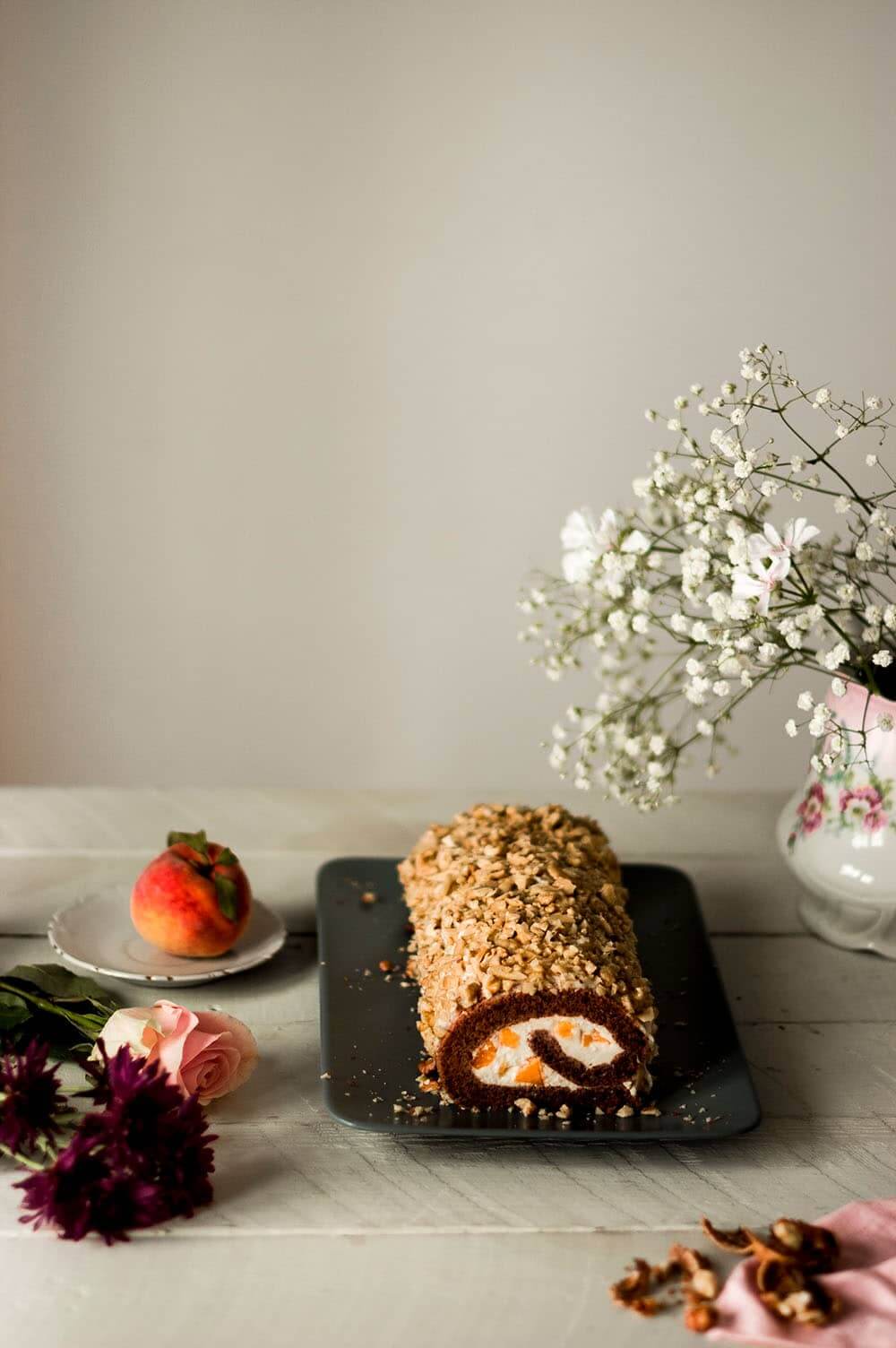 En una mesa de madera blanca hay una una fuente alargada. En ella está el brazo de gitano de nueces y crema de chocolate blanco y melocotón. Alrededor hay flores, melocotón fresco y flores.