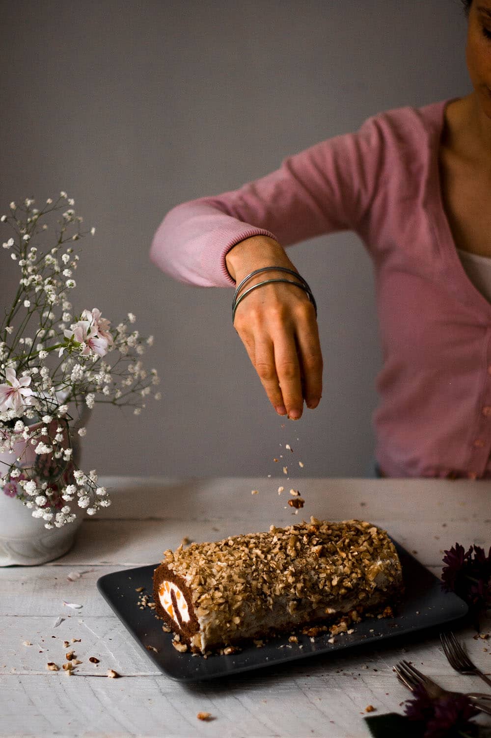 En una mesa de madera blanca hay una una fuente alargada. En ella está el brazo de gitano de nueces y crema de chocolate blanco y melocotón. Una mano está espolvoreando con nueces picadas. 