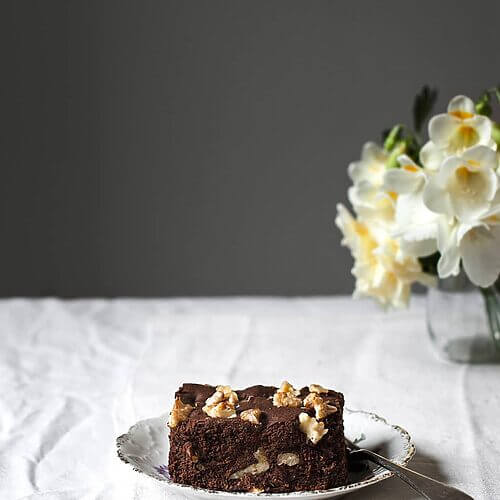 Una mesa cubierta con mantel blanco hay un plato.En el plato hay una ración de brownie casero de chocolate. Está muy jugoso. Alrededor hay flores.