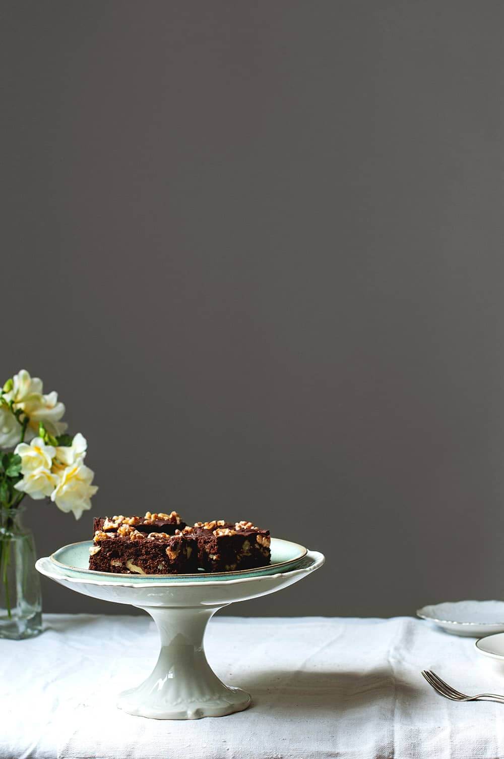 Una mesa cubierta con mantel blanco hay un porta tartas. En él hay varias raciones de brownie esponjoso. Alrededor hay flores