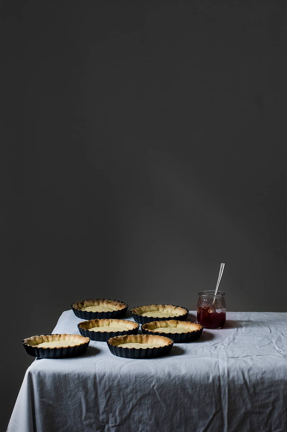 En una mesa cubierta con mantel blanco hay seis tartaletas hechas con pasta sucre original. También se ve un bote de mermelada.