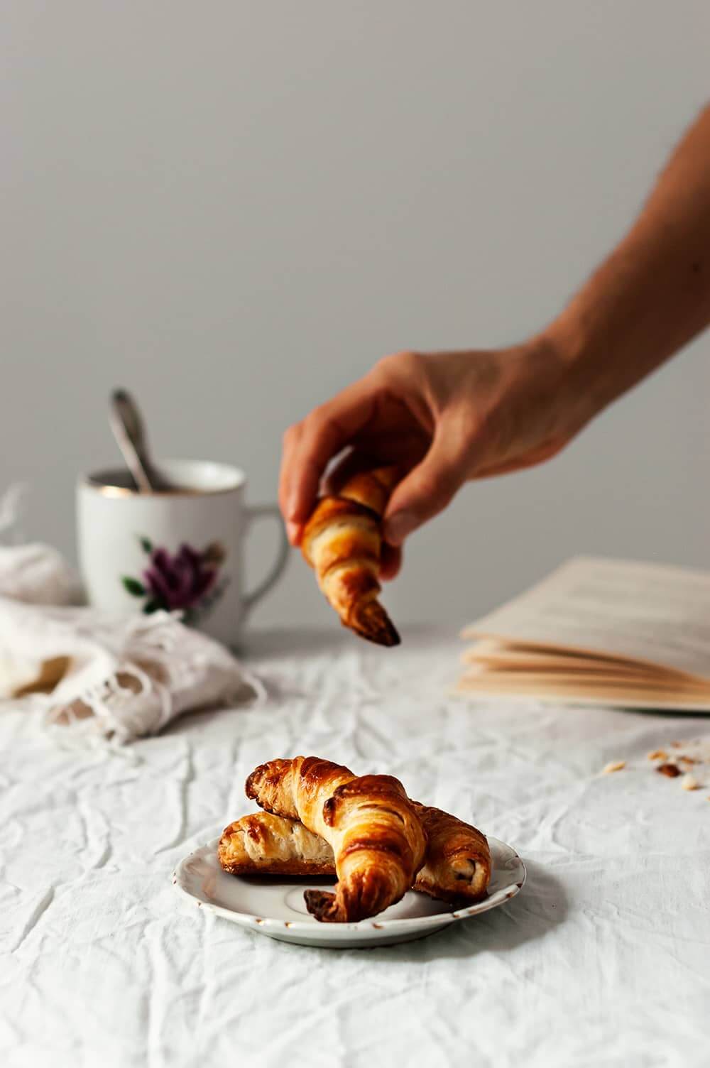 En una mesa cubierta con mantel blanco hay un plato balnco.En el plato hay tres croissants. Una mano está cogiendo uno. Detrás del plato se ven un libro y una taza de té.