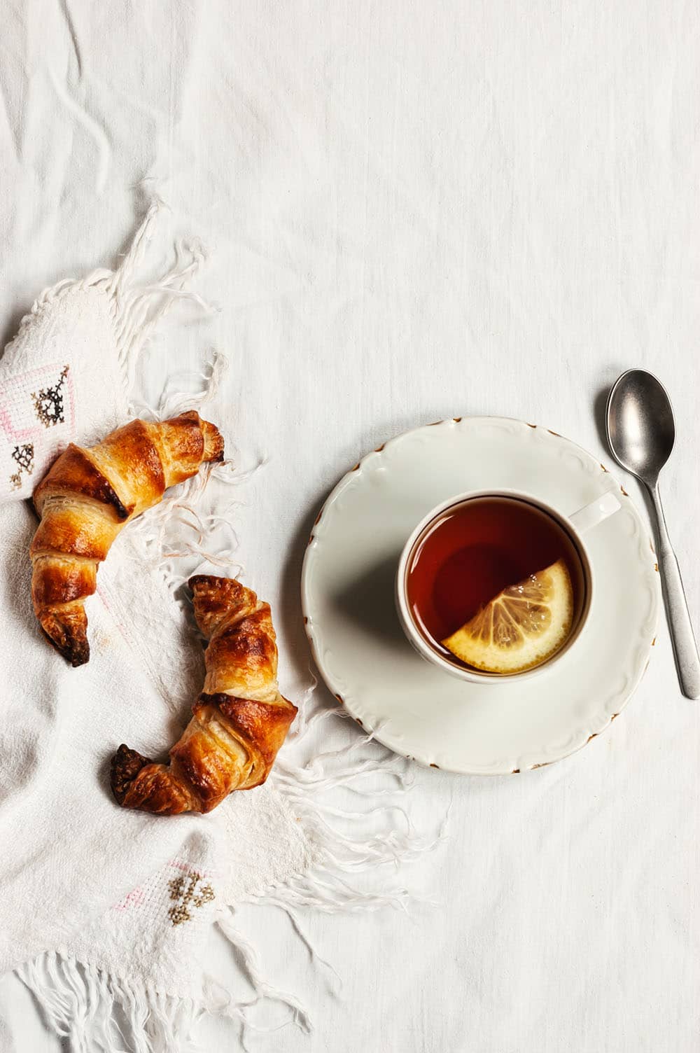 En una mesa cubierta con mantel blanco hay un plato balnco.En el plato una taza de té. Alrededor hay dos croissants.