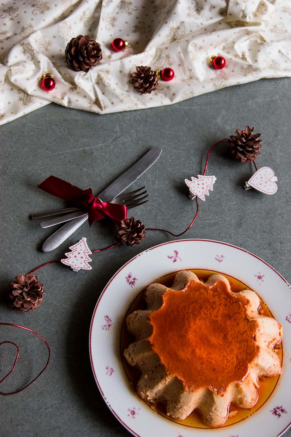 En una mesa de piedra hay un plato. En el medio está el flan de turrón de Jijona. Alrededor hay decoraciones navideñas.