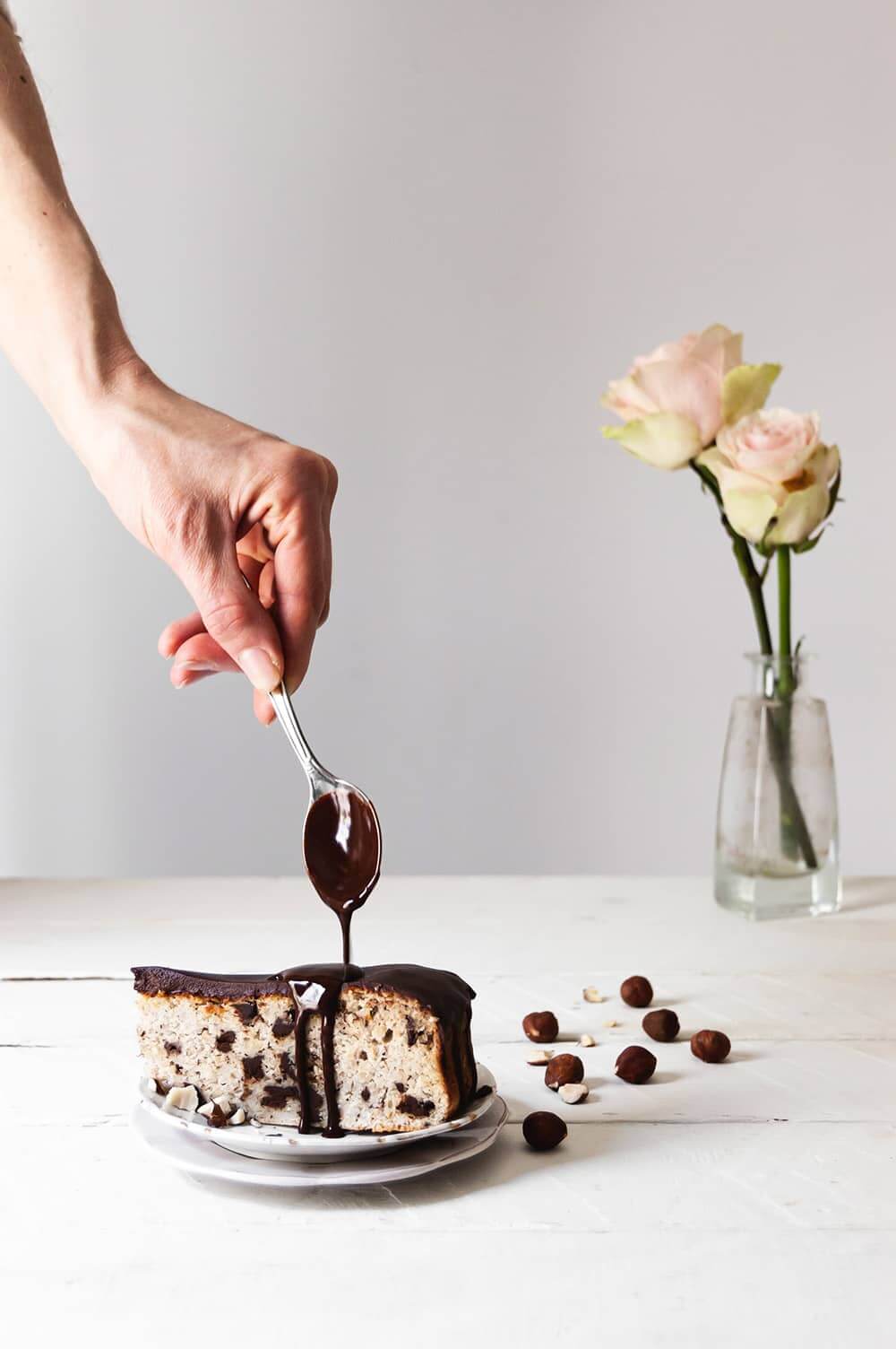 En una mesa de madera blanca hay un plato blanco y en el hay una ración de tarta. Una persona está cubriendo esta tarta con el ganache de chocolate sin nata.