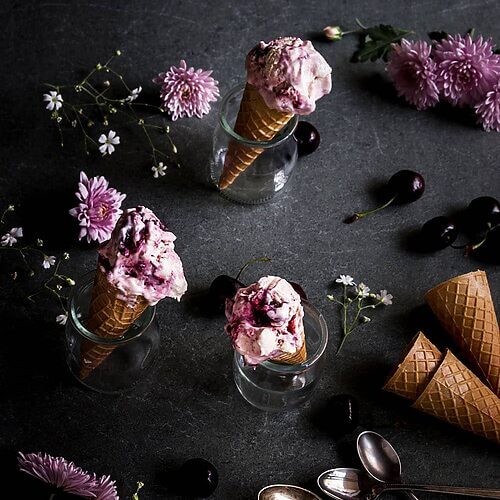 En una mesa de piedra hay tres vasos de cristal. En cada una hay cucuruchos y helado de cheesecake con cerezas sin heladera. Alrededor hay cerezas, flores y cucuruchos vacíos.