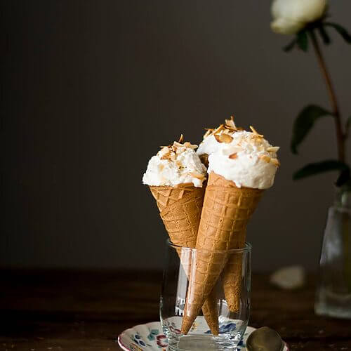 En una mesa de madera hay un plato. En el plato hay un vaso de cristal con dos cucuruchos. Cada una está llena de helado de coco casero. Detrás hay una flor blanca.