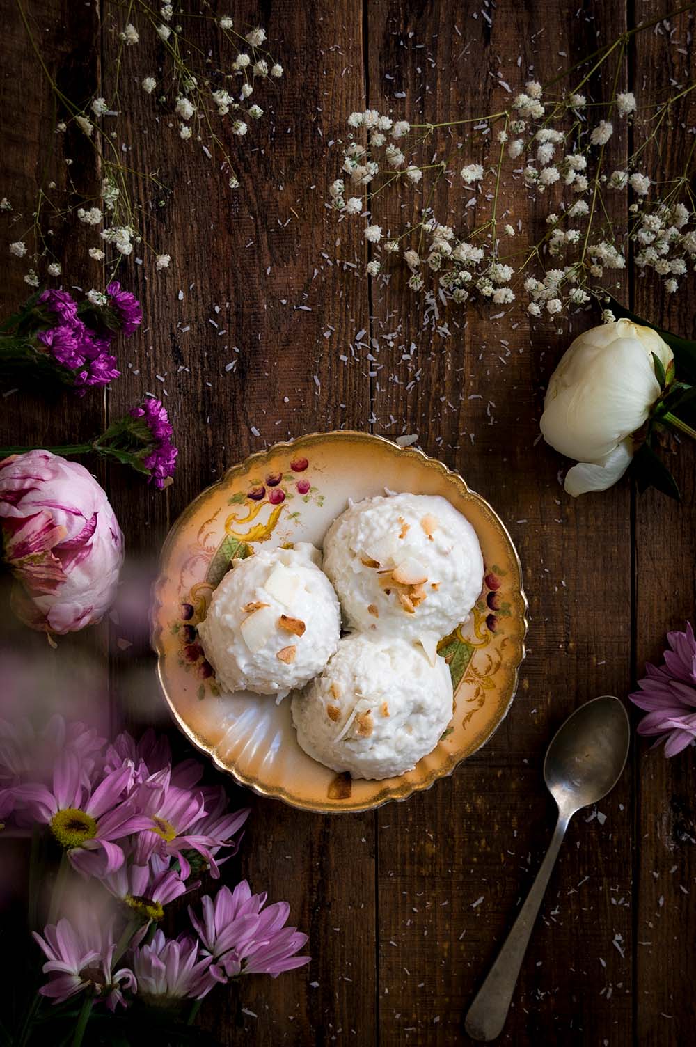 En una mesa de madera hay un plato lleno de helado de coco casero cremoso. Alrededor hay pétalos, flores y cubiertos.