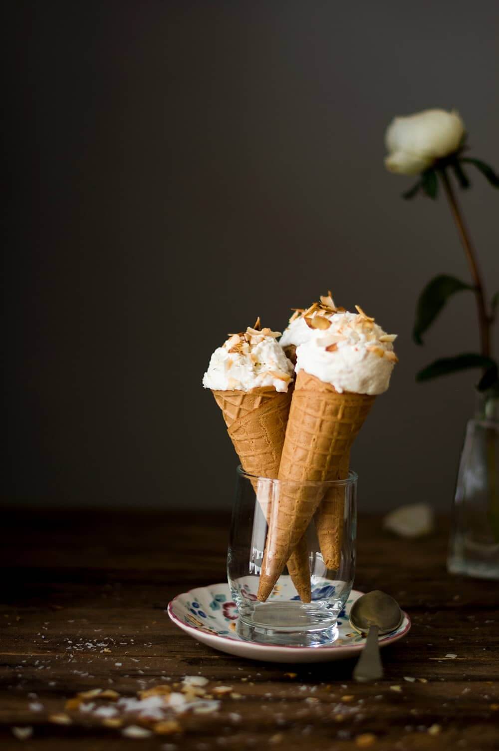 En una mesa de madera hay un plato. En el plato hay un vaso de cristal con dos cucuruchos. Cada una está llena de helado de coco casero. Detrás hay una flor blanca.