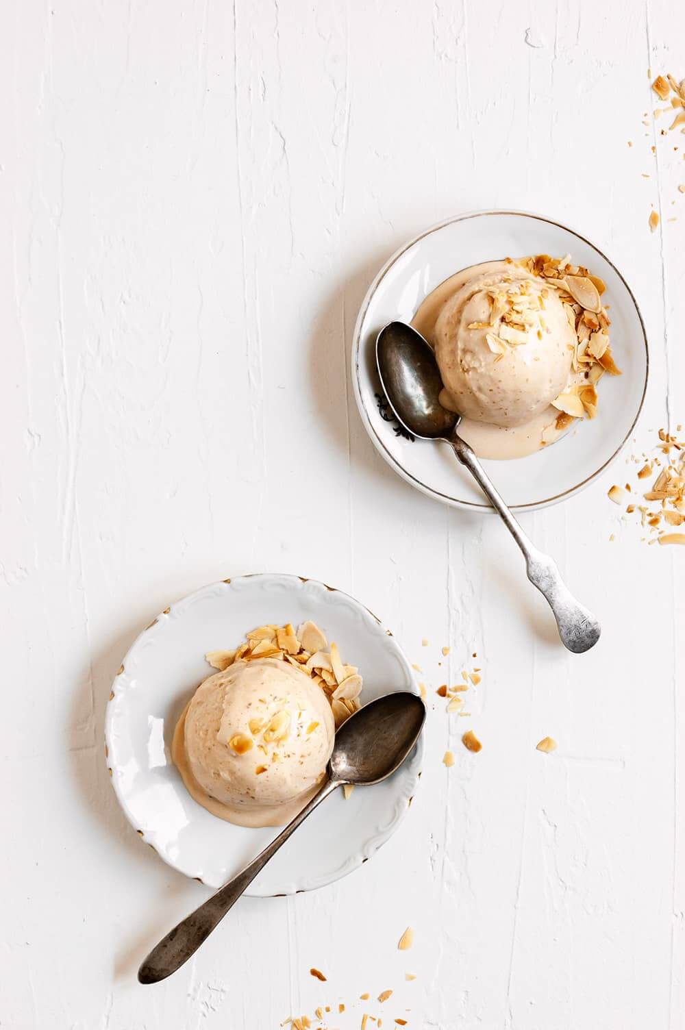 En una mesa de madera blanca hay dos platos. En cada una hay una bola de helado de turrón de Jijona. En cada plato hay una cuchara.