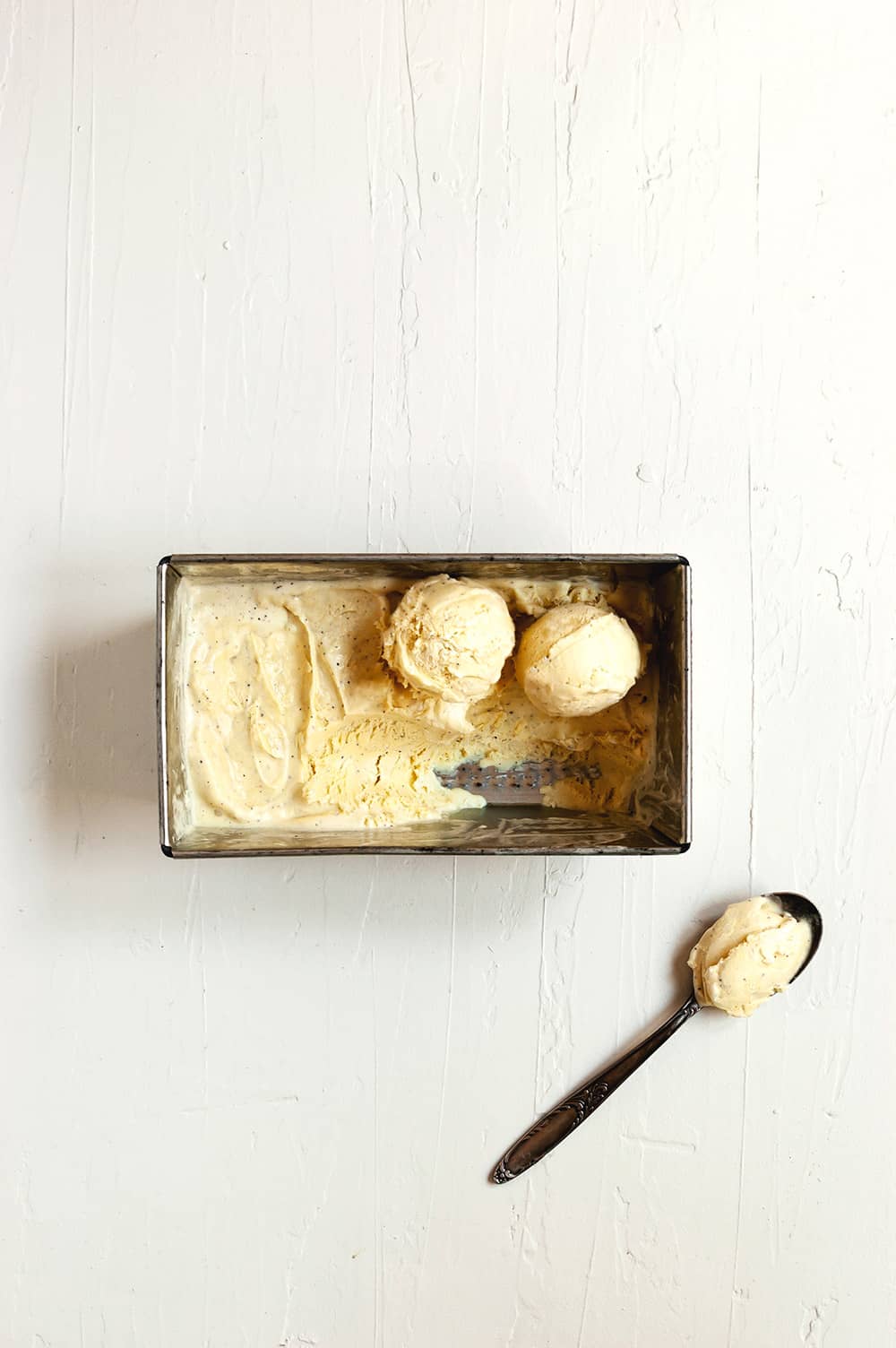 En una mesa de madera blanca hay un recipiente de metal lleno de helado de vainilla casero con huevo. Al lado hay una cuchara con helado en ella.