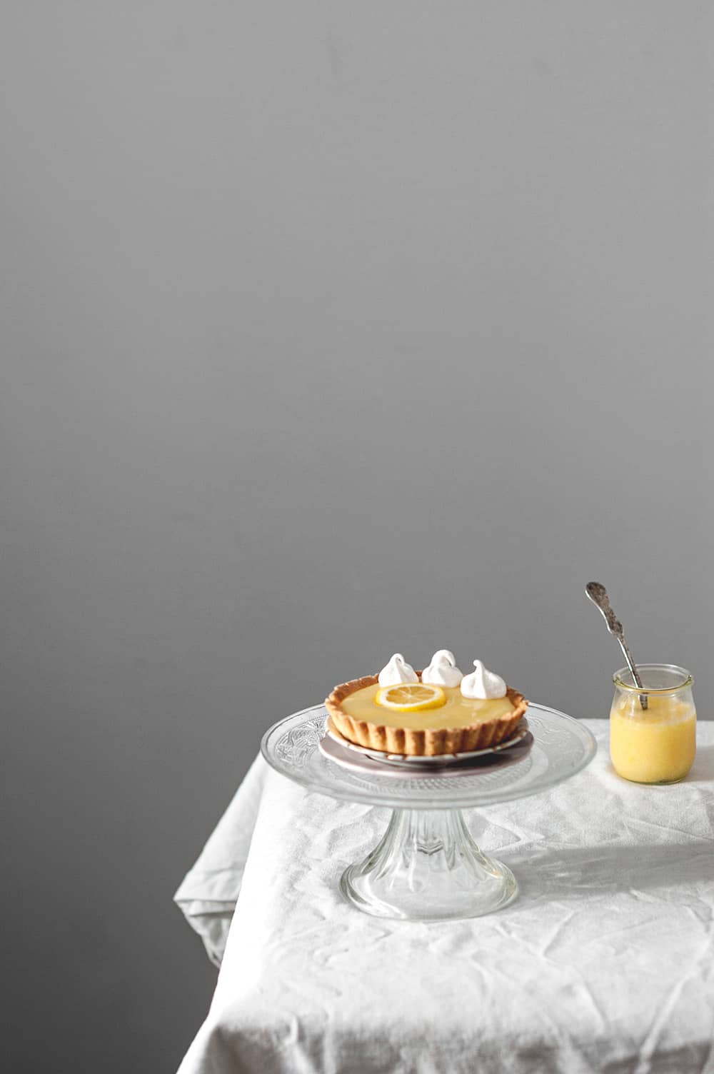 En una mesa cubierta con un mantel blanco hay un porta tartas de cristal. En ella está una tartaleta relleno con Lemon curd casero.