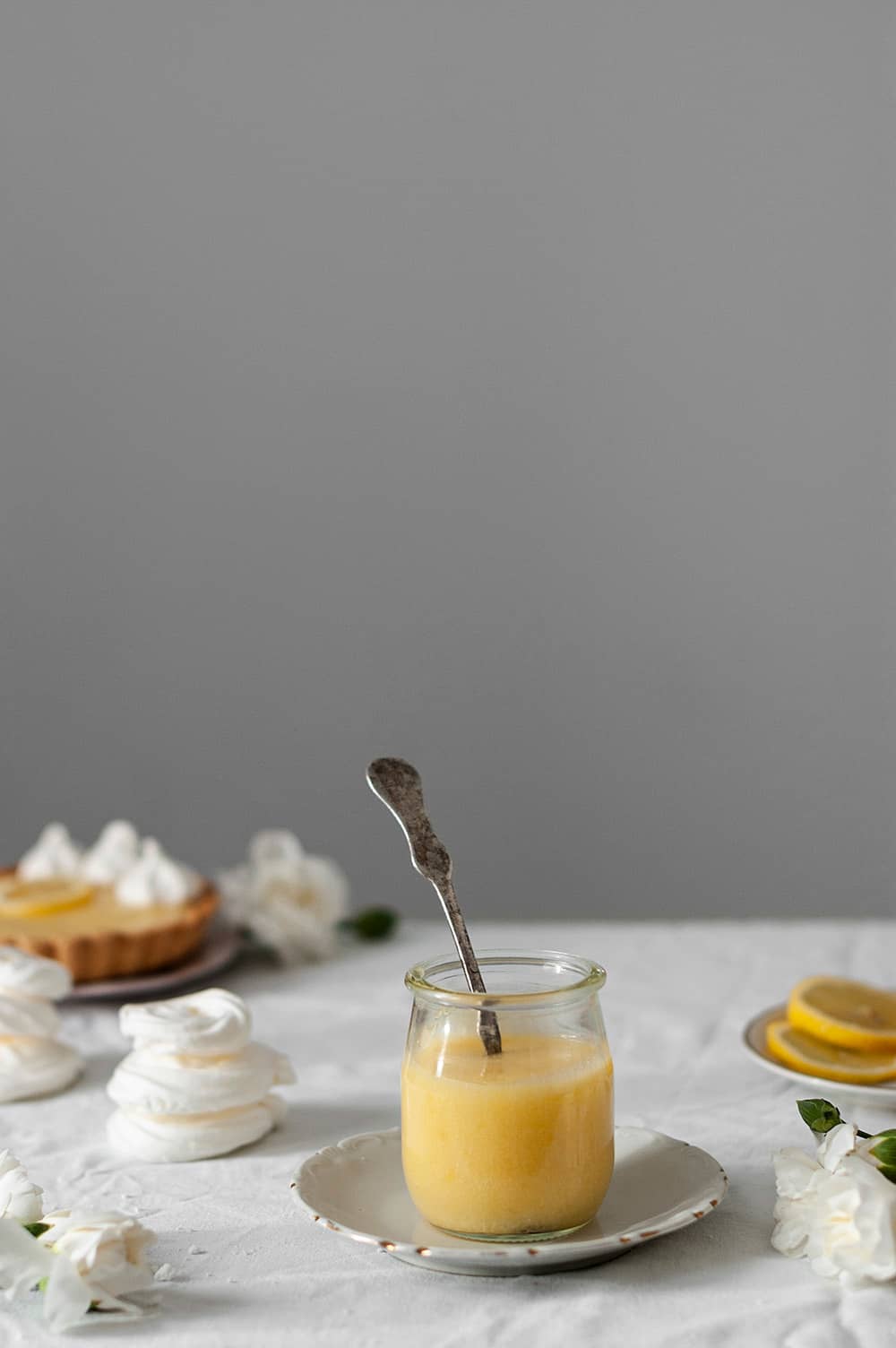 En una mesa cubierta con un mantel blanco hay un plato. En él está un vaso lleno de Lemon curd fácil. En este vaso hay una cuchara. Alrededor hay tartaletas y merengues. 