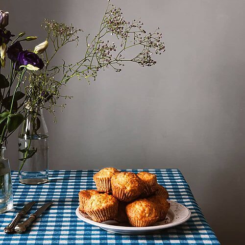 En una mesa cubierta con un mantel de cuadros hay un plato lleno de magdalenas de manzana y coco rallado. Alrededor hay cuchillos y flores.