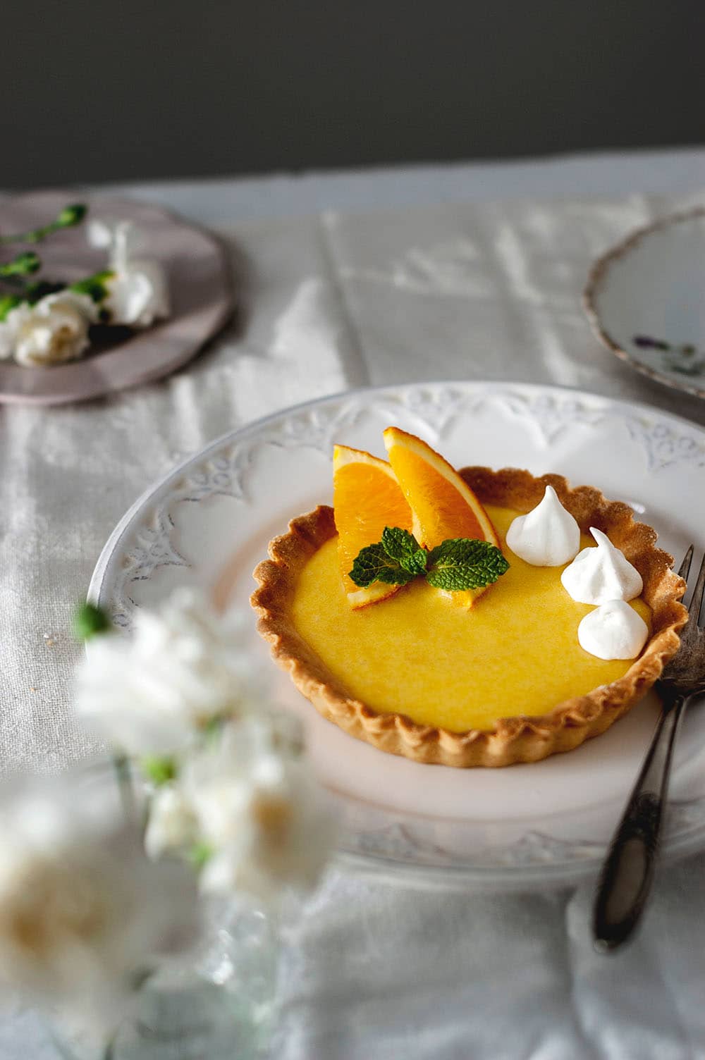 En una mesa cubierta con un mantel blanco hay un plato blanco grande . En el hay otro plato con una tartaleta llena de Orange curd fácil y rápido. Está decorada con merengues duros. Alrededor hay flores. 