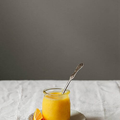 En una mesa cubierta con un mantel blanco hay un plato. En él está un vaso lleno de Orange curd inglés. En este vaso hay una cuchara. Alrededor hay trozos de naranja. 