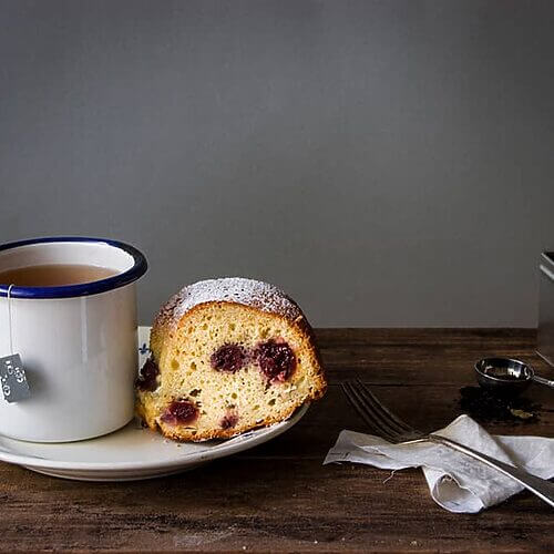 En una mesa de madera hay un plato con una ración de plum cake de guindas esponjoso. Al lado del plato hay una taza de té.