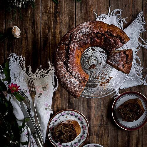 En una mesa de madera un porta tartas de cristal. En él está el plum cake de pasas esponjoso. Ya está cortada. Alrededor hay dos raciones de este bizcocho, flores y servilletas de textil.