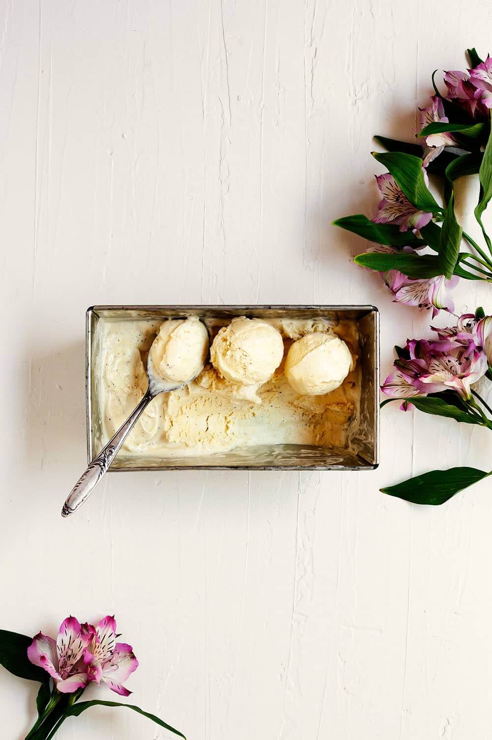 En una mesa de madera blanca hay un recipiente de metal lleno de helado de vainilla casero. Al lado hay una cuchara con helado en ella. Alrededor hay flores frescas.