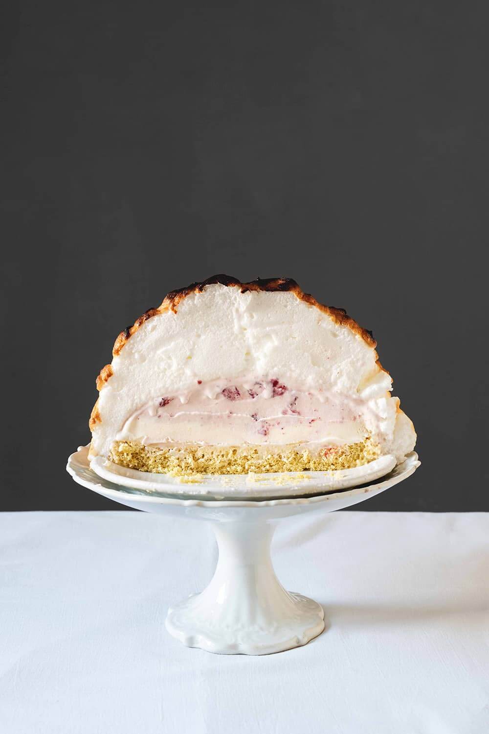 Baked Alaska Una mesa está cubierta con un mantel blanco. En la mesa hay una porta tartas vintage. Encima está el Baked Alaska cortado en la mitad.
