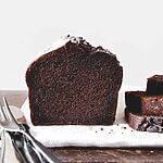 Una mesa está cubierta de madera. Encima hay un paño de cocina blanco. Encima está el bizcocho de chocolate y calabacín, Se ve muy esponjoso. Al lado hay un cuchillo y un par de rebanadas de bizcocho.