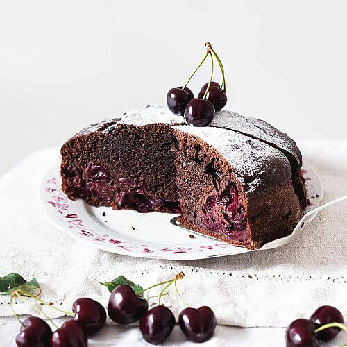 En una mesa cubierta con un mantel blanco. encima está un plato blanco. En el plato está el bizcocho de cerezas y chocolate. Ya está cortada. Se ve muy esponjoso. Alrededor hay cerezas.